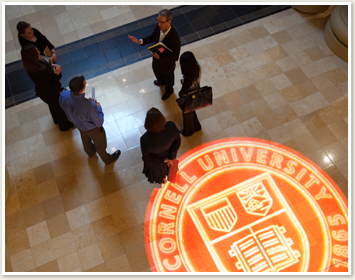 Aerial view of students and teacher standing and talking near Cornell seal on floor